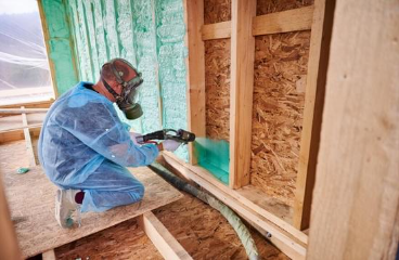 Construction worker insulating wooden frame house with polyurethane foam using plural component gun.